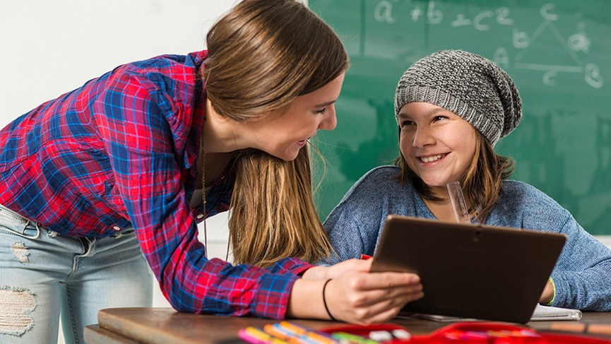 Lehrkraft und Mädchen mit Tablet, im Hintergrund eine Tafel mit mathematischer Formel